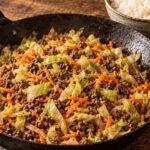Skillet of ground beef, cabbage and carrots with a bowl of rice on a wooden table